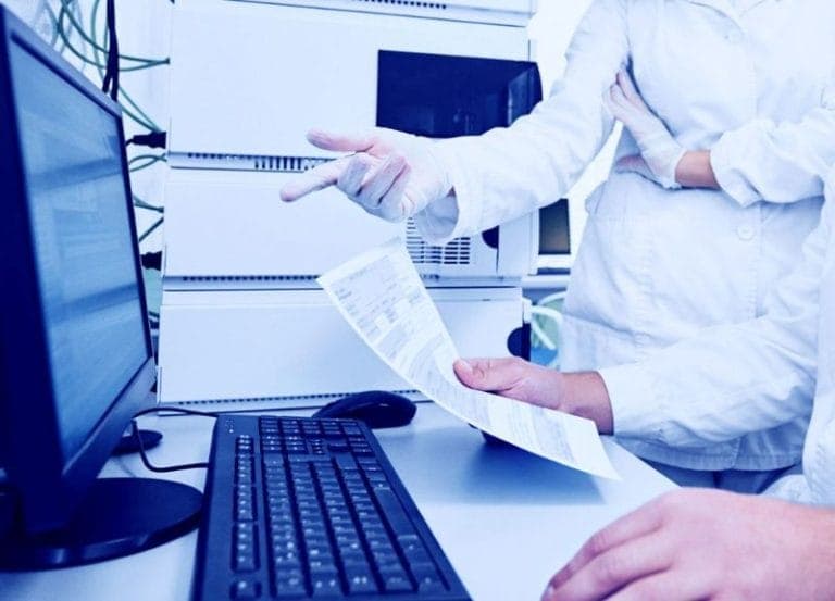 Laboratório farmacêutico de alta tecnologia com cientistas discutindo dados em um grande monitor, equipamentos de segurança e reagentes químicos.
